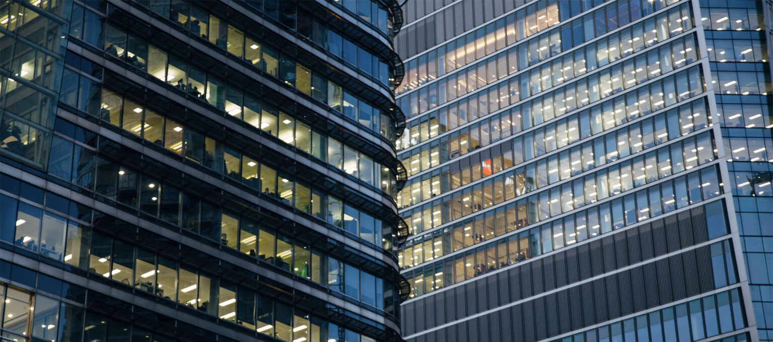 Two modern glass office buildings with illuminated windows, showing multiple floors and offices at twilight.