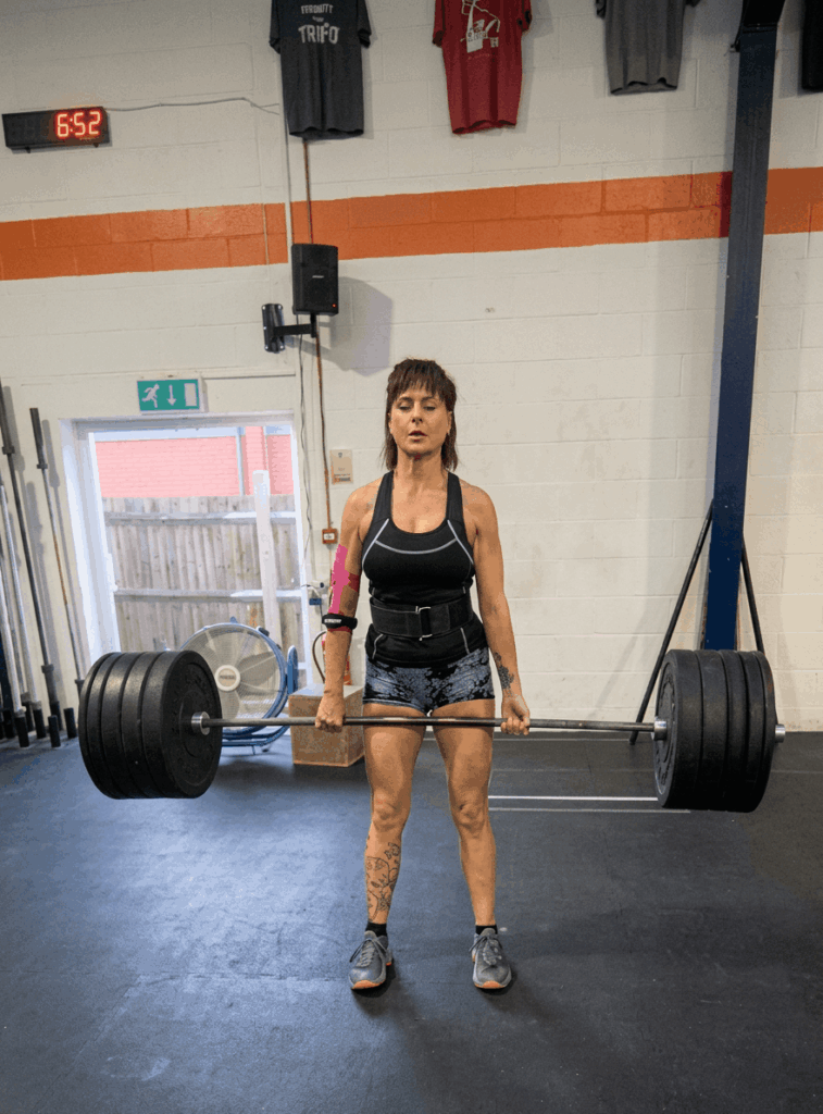 Person lifting a heavy barbell in a gym, wearing athletic clothes and shoes. There are shirts and a clock on the wall, gym equipment around, and the person has tattoos on their leg and an arm sleeve on their right arm.