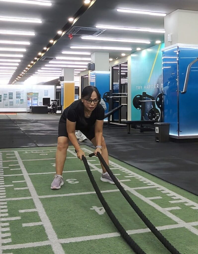 A woman in athletic wear exercises with battle ropes on a turf field inside a modern gym, surrounded by workout equipment and motivational wall art.