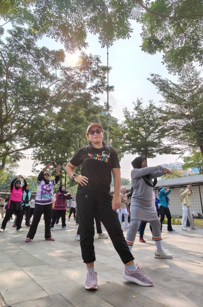 A woman in sunglasses poses confidently in the foreground, while a group of people do aerobic exercises in a park under trees on a sunny day.