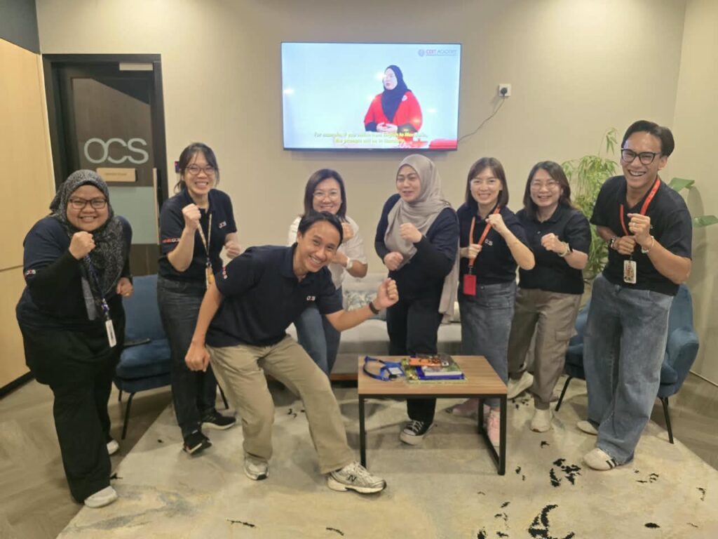 A group of eight smiling people pose enthusiastically in an office, some making playful fist gestures. They wear lanyards and casual attire. A TV in the background shows a woman in a hijab speaking.