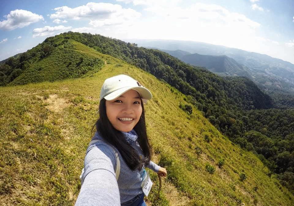 A woman wearing a cap smiles while taking a selfie on a grassy hilltop with green mountains and a blue sky in the background.