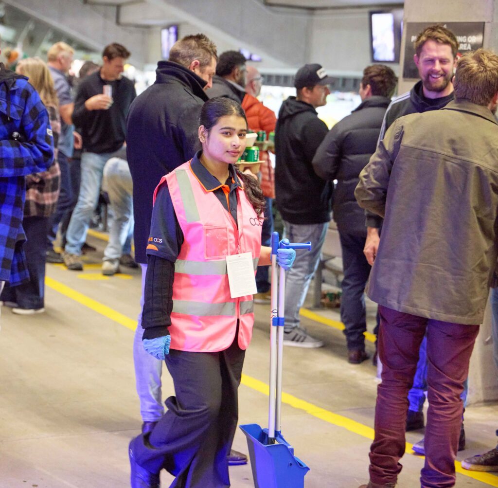 A staff member in a pink high-visibility vest and blue gloves holds a dustpan and brush whilst walking through a crowded venue.
