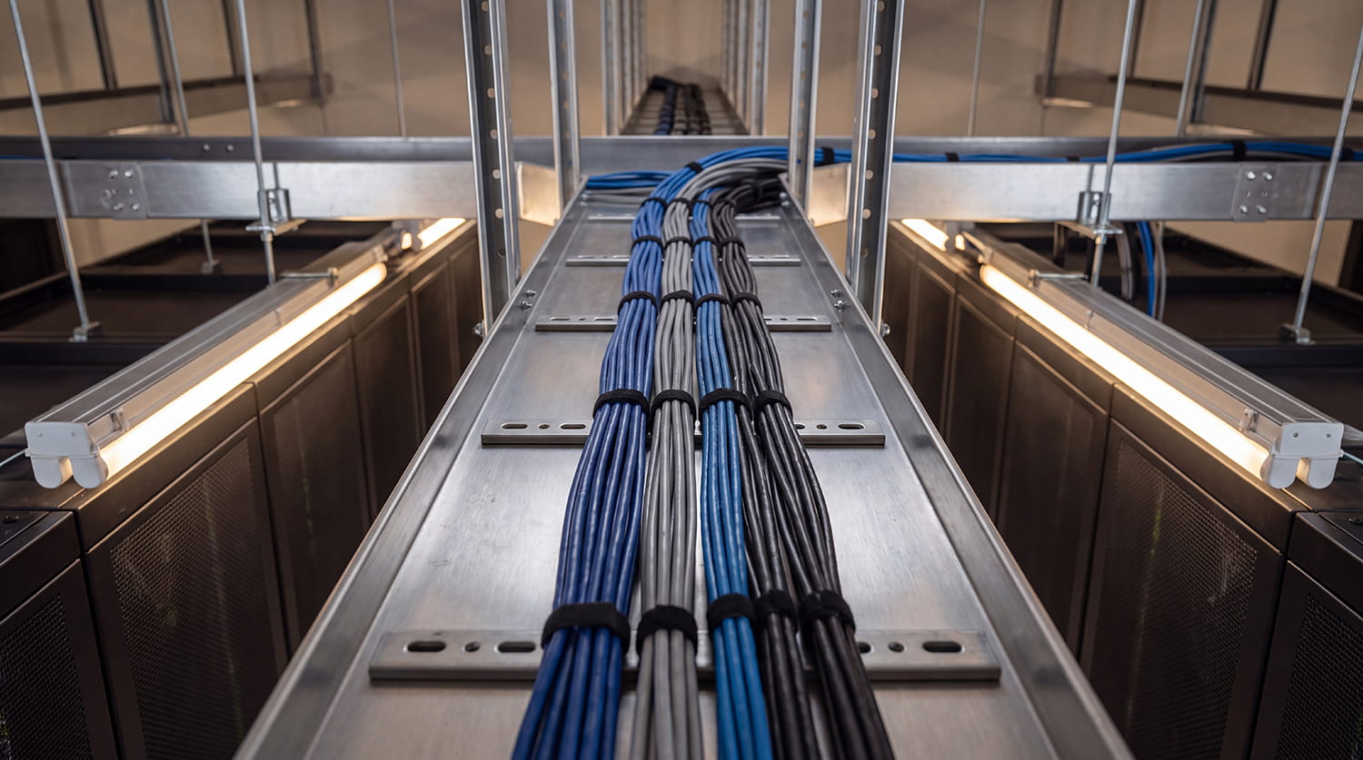 Bundles of blue, grey, and black data cables organised on a metal tray above server racks.