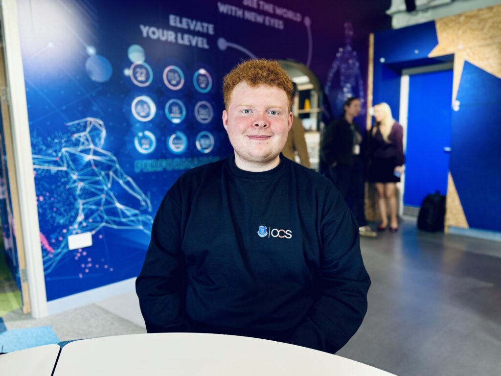 A young man with red hair sits in a workspace with blue walls featuring performance-related graphics and text.