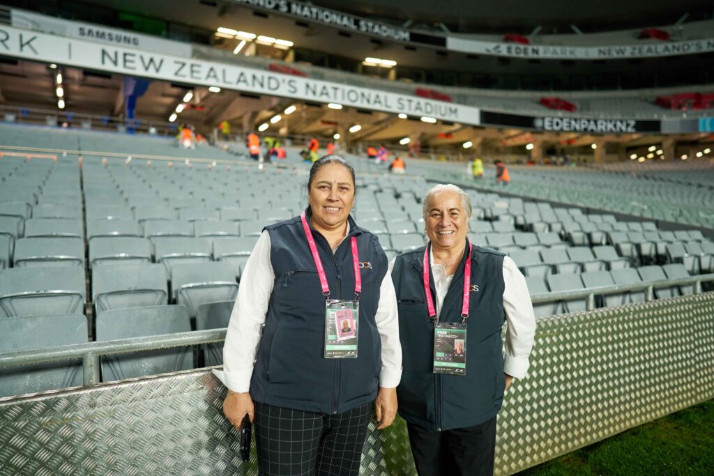 Two smiling women wearing dark waistcoats and event lanyards stand in front of the empty seating rows at Eden Park stadium.