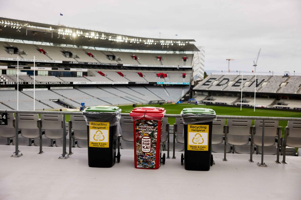 Three recycling bins, two black and one red, lined up in front of an empty stadium seating area.
