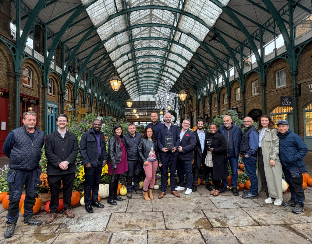 A group of sixteen people stand in a queue for a photograph inside the Covent Garden market hall, with one person in the centre holding a glass award plaque.