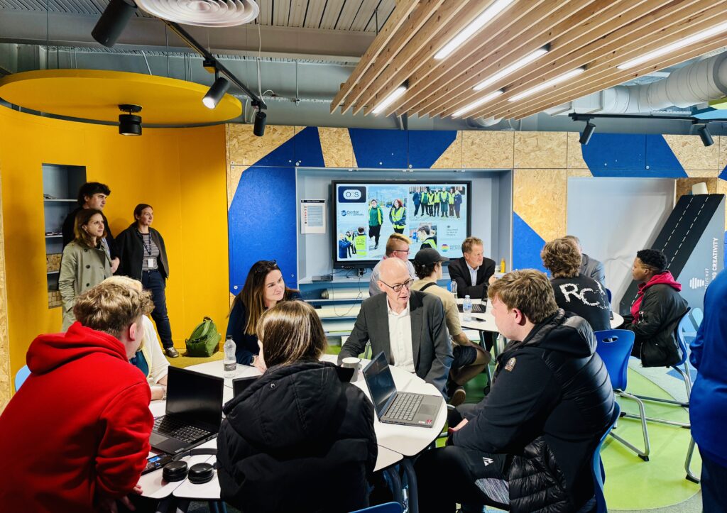 A diverse group of people sitting around a circular table in an office space, with some using laptops whilst a screen in the background displays group photos.