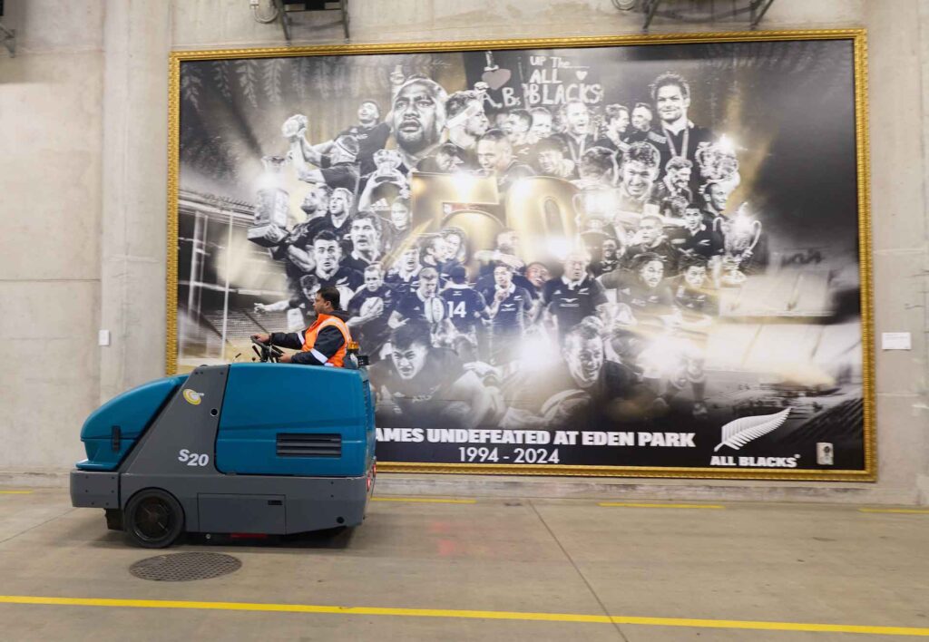 A worker operates a blue floor-cleaning machine in front of a large, gold-framed commemorative poster of the All Blacks rugby team.