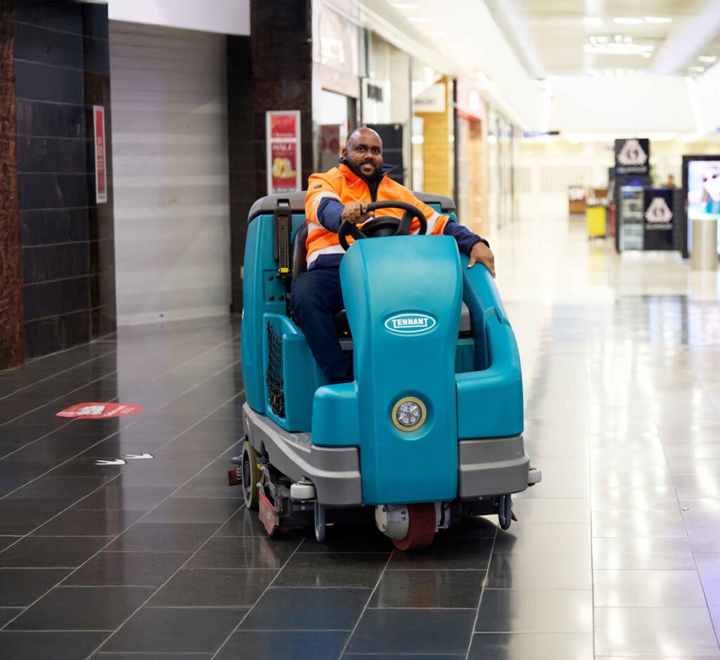 A man in a high-visibility orange jacket drives a teal Tennant floor scrubber through a shopping centre corridor.