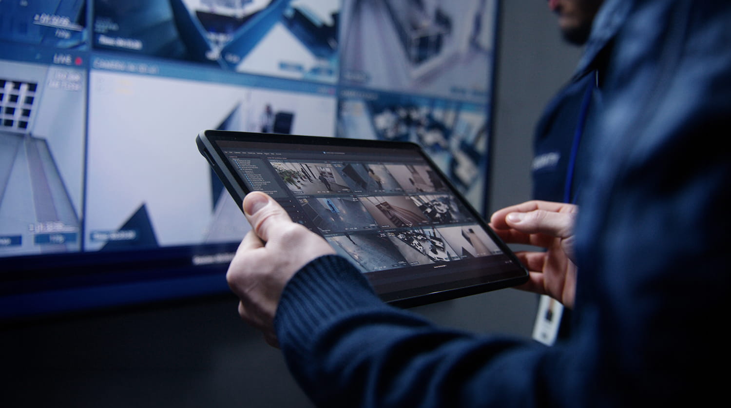 A security guard holding a tablet displaying multiple CCTV feeds, with a large monitoring screen in the background.