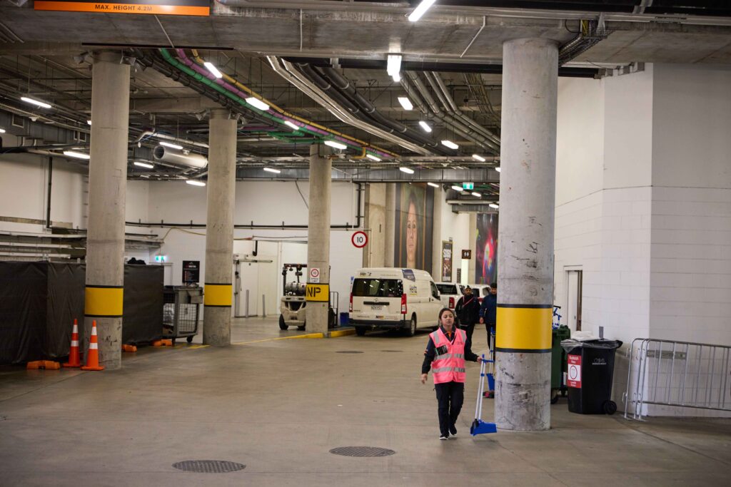 A worker in a pink reflective vest pushes a broom through an industrial loading bay or car park area.